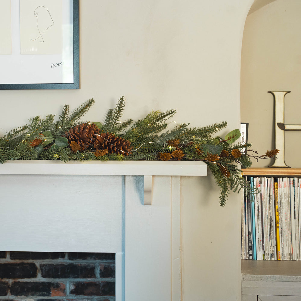 A green pine garland with brown pinecones and warm string lights draped across a white fireplace mantel in a home interior setting.