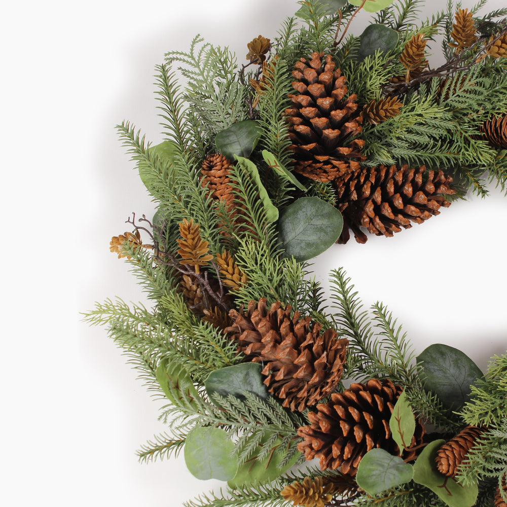 A close up of a festive artificial Christmas wreath made of green pine branches, decorated with large brown pinecones, small orange-brown pinecones, and round green eucalyptus leaves, photographed on a clean white background.