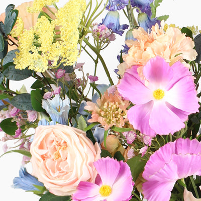 A close-up of a mixed floral arrangement featuring large pink flowers, a pale peach rose, yellow clusters, and soft blue blossoms with green foliage on a white background.