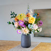 A multicolored floral arrangement featuring soft yellows, purples, pinks, and blues, presented in a matte dark gray ceramic vase on a table runner.