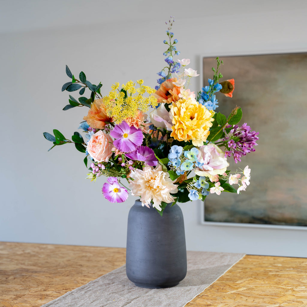 A multicolored floral arrangement featuring soft yellows, purples, pinks, and blues, presented in a matte dark gray ceramic vase on a table runner.