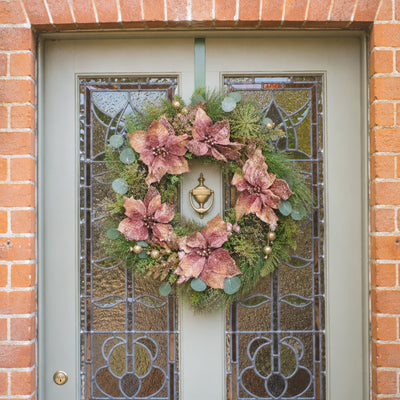 A decorative wreath with pink poinsettia-flowers, green foliage, and eucalyptus hangs on a gray front door with stained glass panels, set against a red brick wall.