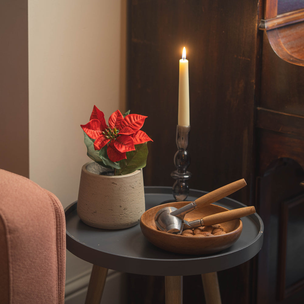 A cozy indoor scene with a small round gray side table holding a potted red poinsettia, a tall lit candle and a wooden bowl of walnuts and nutcrackers. Part of a pink upholstered chair is visible at left, and a piano.