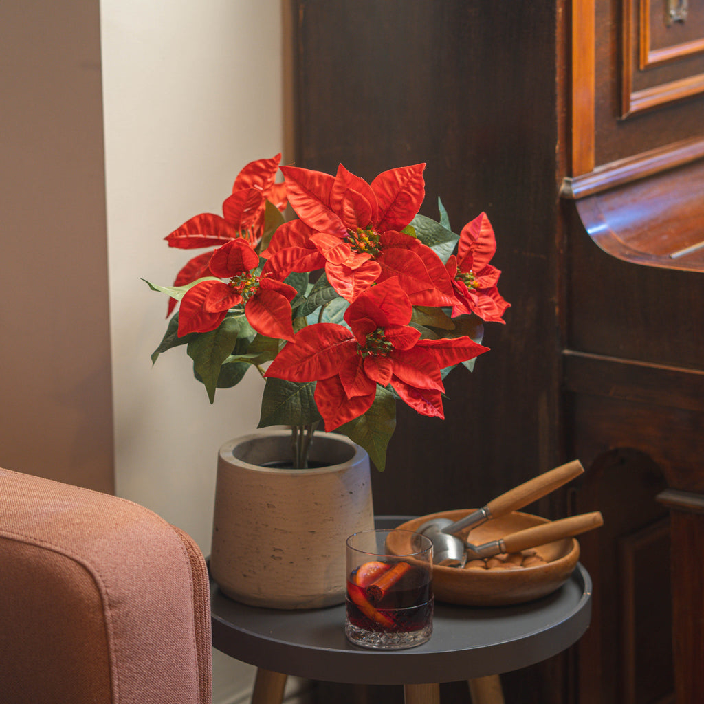 A red artificial poinsettia plant in a grey textured pot, styled on a dark gray side table with a glass and a wooden bowl of walnuts next to a piano.