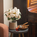 White artificial poinsettia plant in a grey textured pot, styled on a dark gray side table next to a glass and a wooden bowl of walnuts in a cozy room setting.