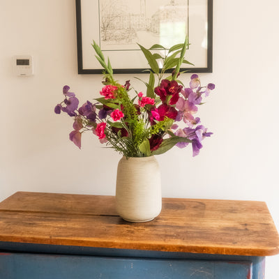 A textured cream ceramic vase filled with a colorful floral arrangement of pink, purple, and red flowers with green leaves, sitting on a rustic wooden table against a white wall.