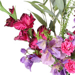 A close up of a mixed floral arrangement featuring deep magenta gladiolus, purple sweet peas, bright pink carnations, and eucalyptus and foliage on a plain white background.