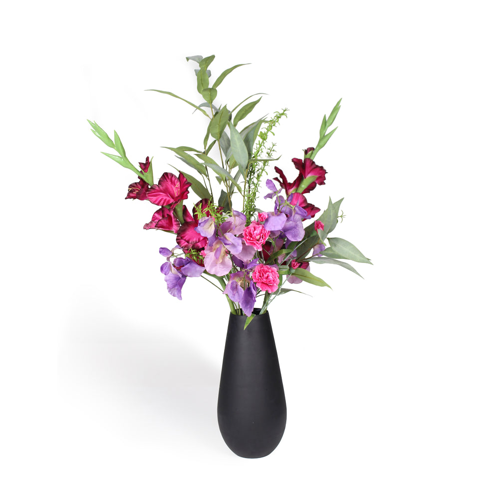 A black ceramic vase filled with a floral arrangement featuring pink and purple flowers with green leaves, set against a plain white background.