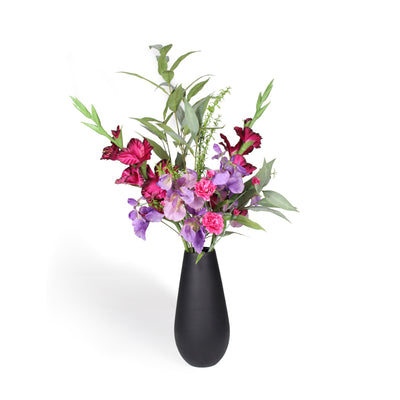A black ceramic vase filled with a floral arrangement featuring pink and purple flowers with green leaves, set against a plain white background.