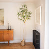 A tall artificial tree with thin green leaves and a light beige trunk, styled in a woven basket pot on a wooden floor in a room corner with a mid-century sideboard, framed art, and a radiator.