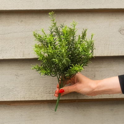 A hand with red nail polish holds a small green artificial rosemary stem against a light gray siding background.