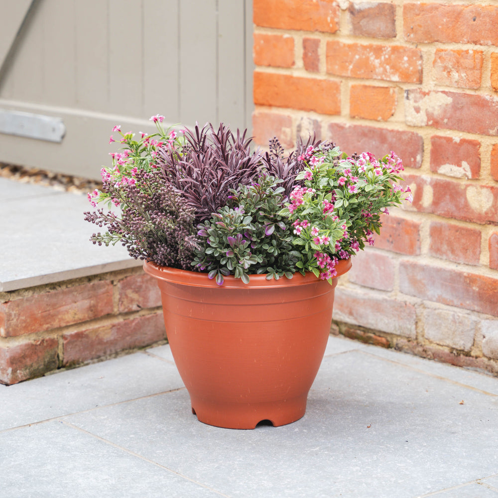Plastic pot with flowering purple and pick plants with foliage against a brick wall.