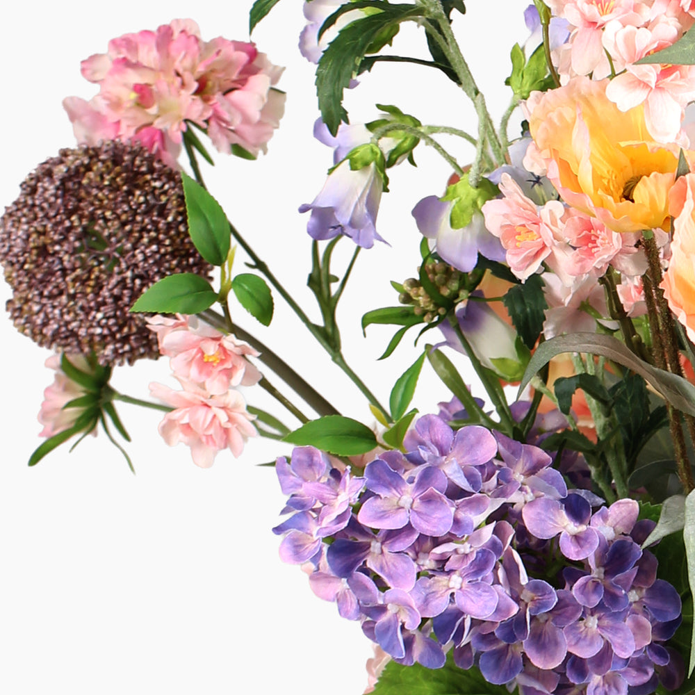 A close-up of a mixed floral arrangement featuring purple, pink and peach flowers, green leaves, and a round seed head on a plain white background.