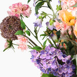 A close-up of a mixed floral arrangement featuring purple, pink and peach flowers, green leaves, and a round seed head on a plain white background.