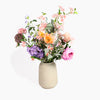 A mixed artificial flower bouquet in a textured beige ceramic vase, featuring pink, orange, purple, and white blossoms with green leaves, set against a plain white background.