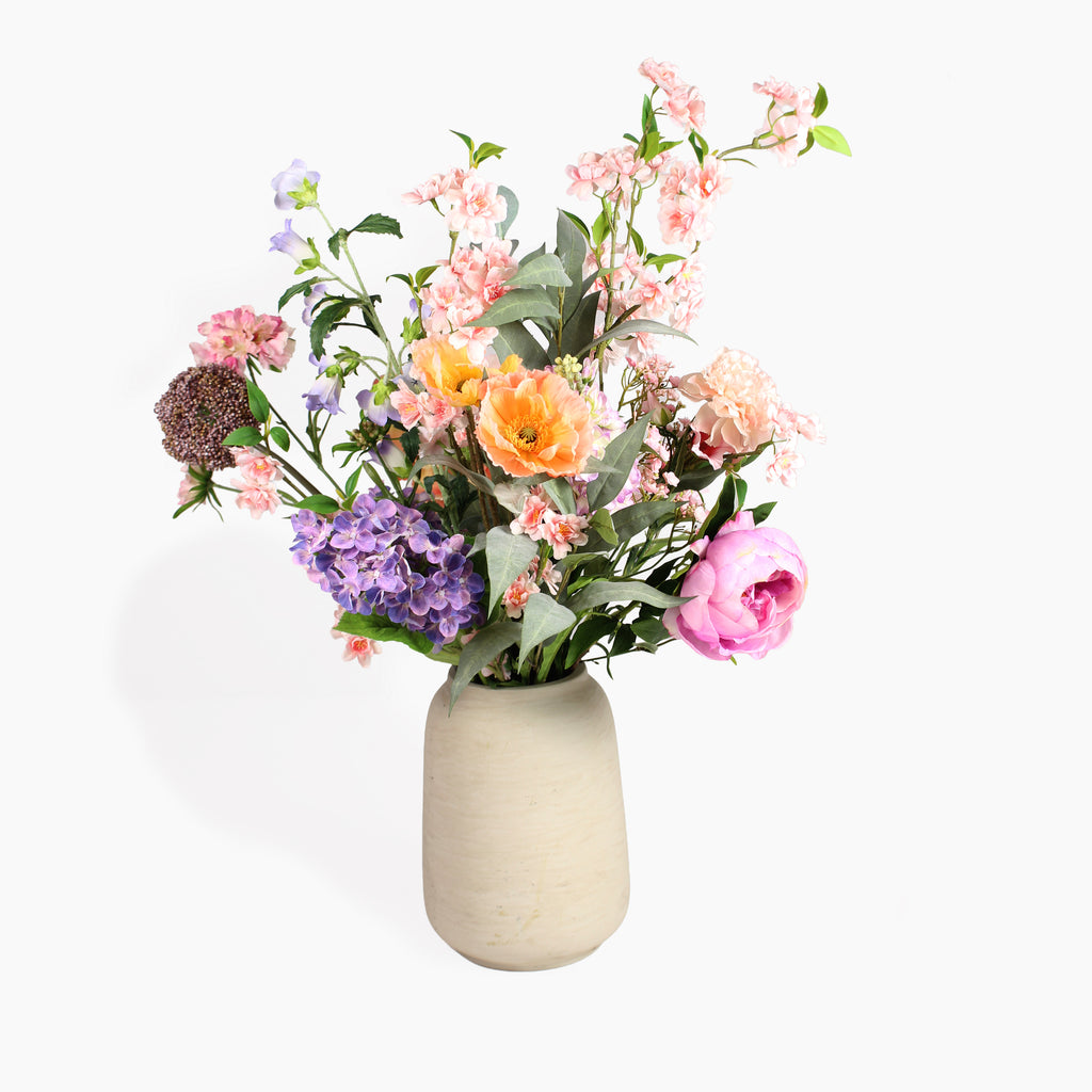 A mixed artificial flower bouquet in a textured beige ceramic vase, featuring pink, orange, purple, and white blossoms with green leaves, set against a plain white background.