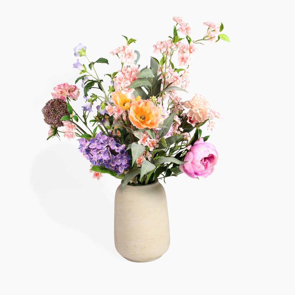 A mixed artificial flower bouquet in a textured beige ceramic vase, featuring pink, orange, purple, and white blossoms with green leaves, set against a plain white background.