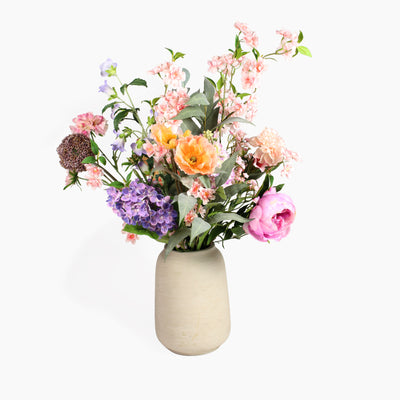 A mixed artificial flower bouquet with tall trailing blossom and a mixture of large and small flower soft coloured flowers in a textured beige ceramic vase, set against a white background.