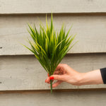 A hand with red nail polish holds a small artificial green grass-like plant in front of a light gray horizontal siding wall.