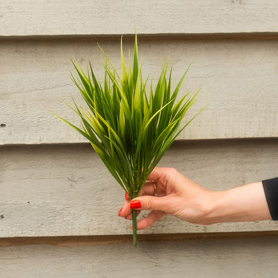 A hand with red nail polish holds a small artificial green grass-like plant in front of a light gray horizontal siding wall.