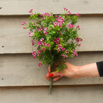 A hand with red nail polish holds a bouquet of artificial pink and white flowers with green leaves against a beige wooden siding background.