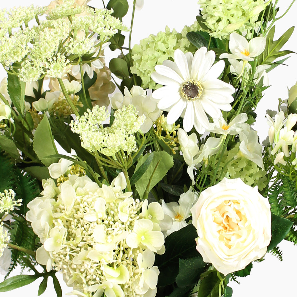 A close-up of a white and cream floral arrangement with green foliage on a white background.