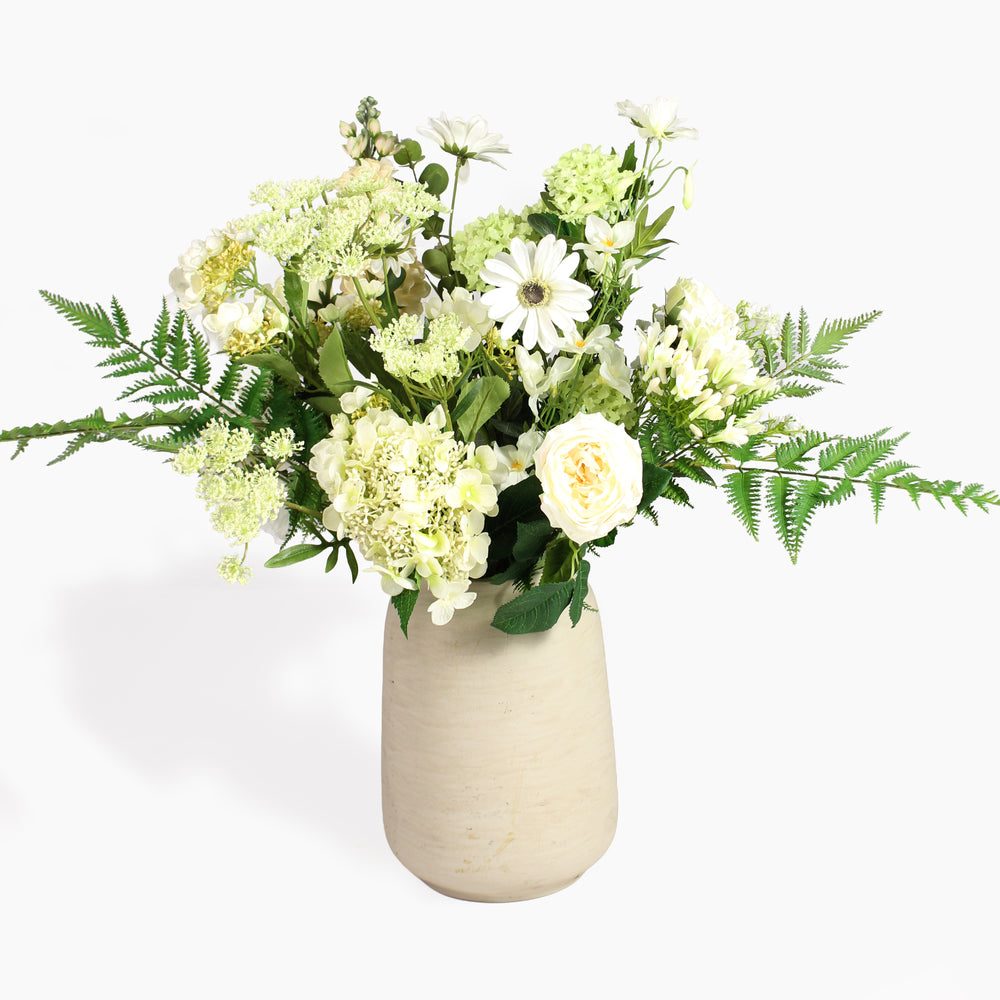 An artificial floral arrangement featuring white and cream flowers, green foliage, and ferns, presented in a textured beige ceramic vase against a clean white background.