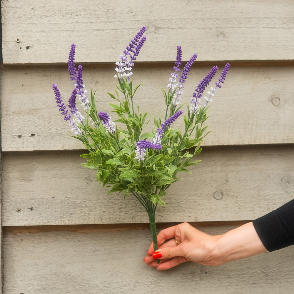 An artificial lavender bouquet with purple and white flowers and green leaves, held by a hand against a gray wooden siding background.