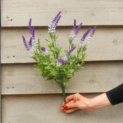 An artificial lavender bouquet with purple and white flowers and green leaves, held by a hand against a gray wooden siding background.