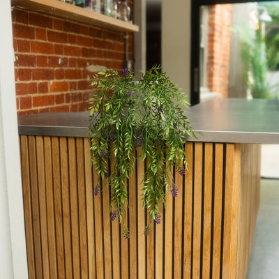 A green artificial hanging plant with small purple flowers draped over a modern wooden slat counter in an indoor setting.