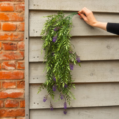 A person's hand with red nail polish holds a bunch of artificial green leaves and purple wisteria flowers against a gray wooden siding wall next to a red brick wall.