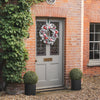 A festive wreath with red ornaments and white faux snow hangs on a gray front door set in a red brick house, ivy growing up the left with potted boxwood plants on either side.