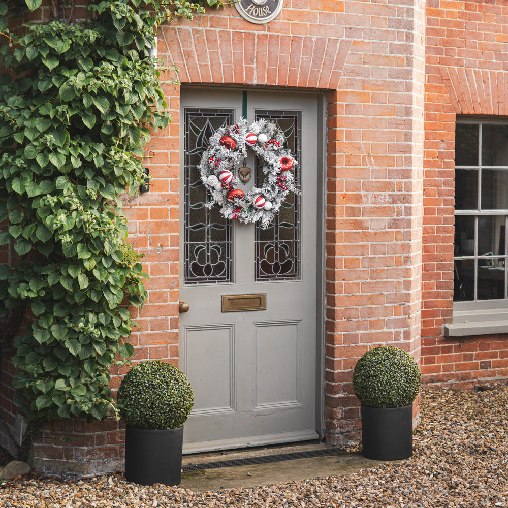 A festive wreath with red ornaments and white faux snow hangs on a gray front door set in a red brick house, ivy growing up the left with potted boxwood plants on either side.