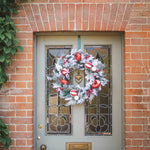 A festive Christmas wreath with white faux snow, red ornaments, and berries hangs on a gray front door with stained glass panels, set against a red brick wall.