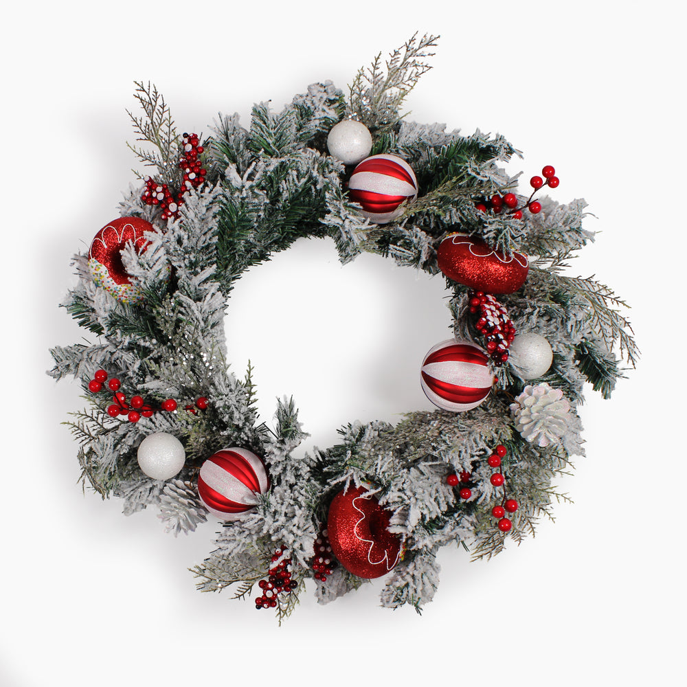A festive Christmas wreath with frosted green pine branches, red berries, and red and white striped doughnut ornaments, photographed against a plain white background.