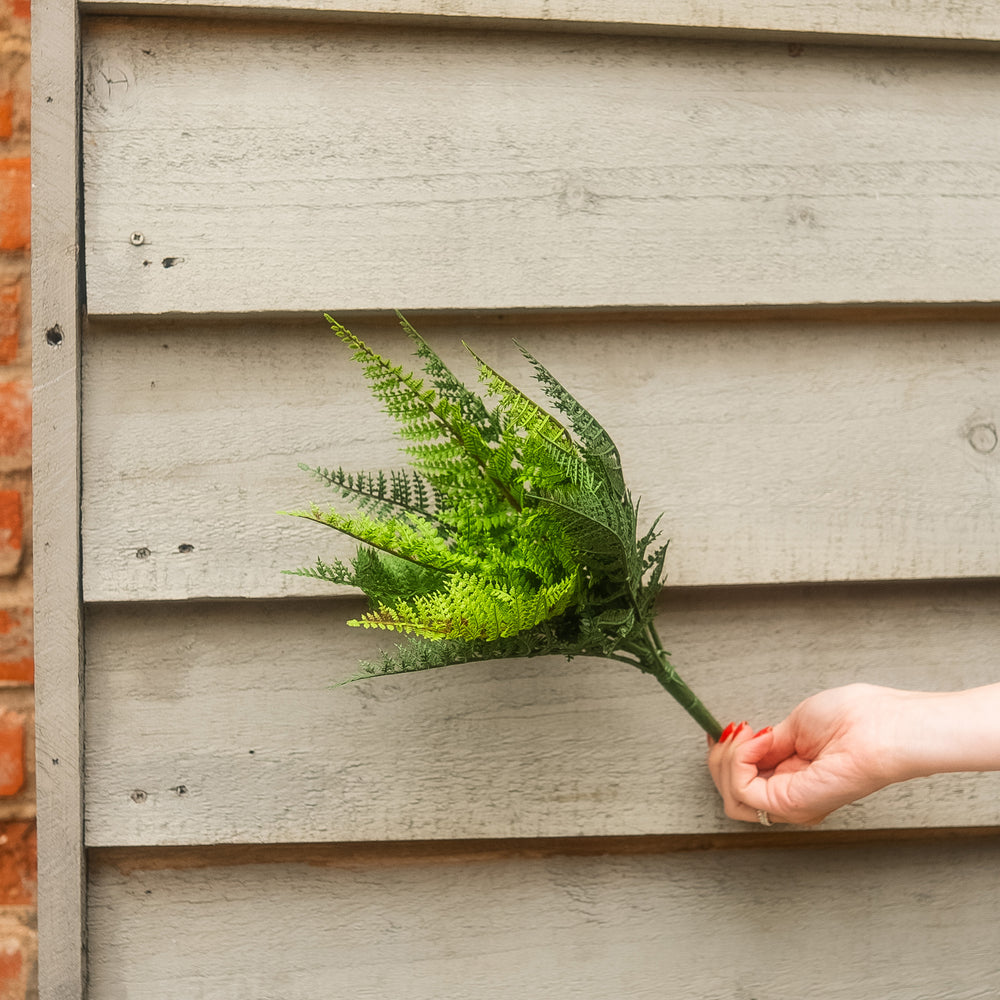 A hand with red nail polish holds a green artificial fern bouquet against a light gray wooden siding background.