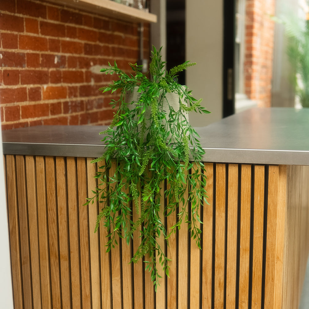 A green trailing artificial plant in a clear glass pot, styled on the edge of a modern wooden slat counter with a metal top.