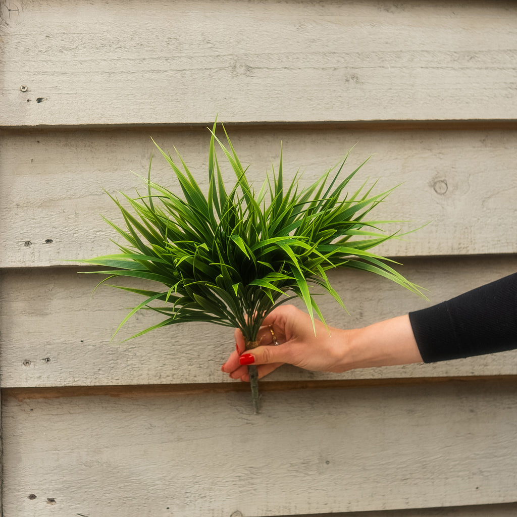 A hand with red nail polish holds a small artificial green plant with long, slender leaves against a light gray wooden plank background.