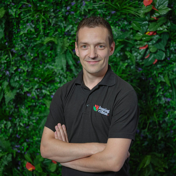 Man wearing a black shirt with a logo standing in front of a green leafy background