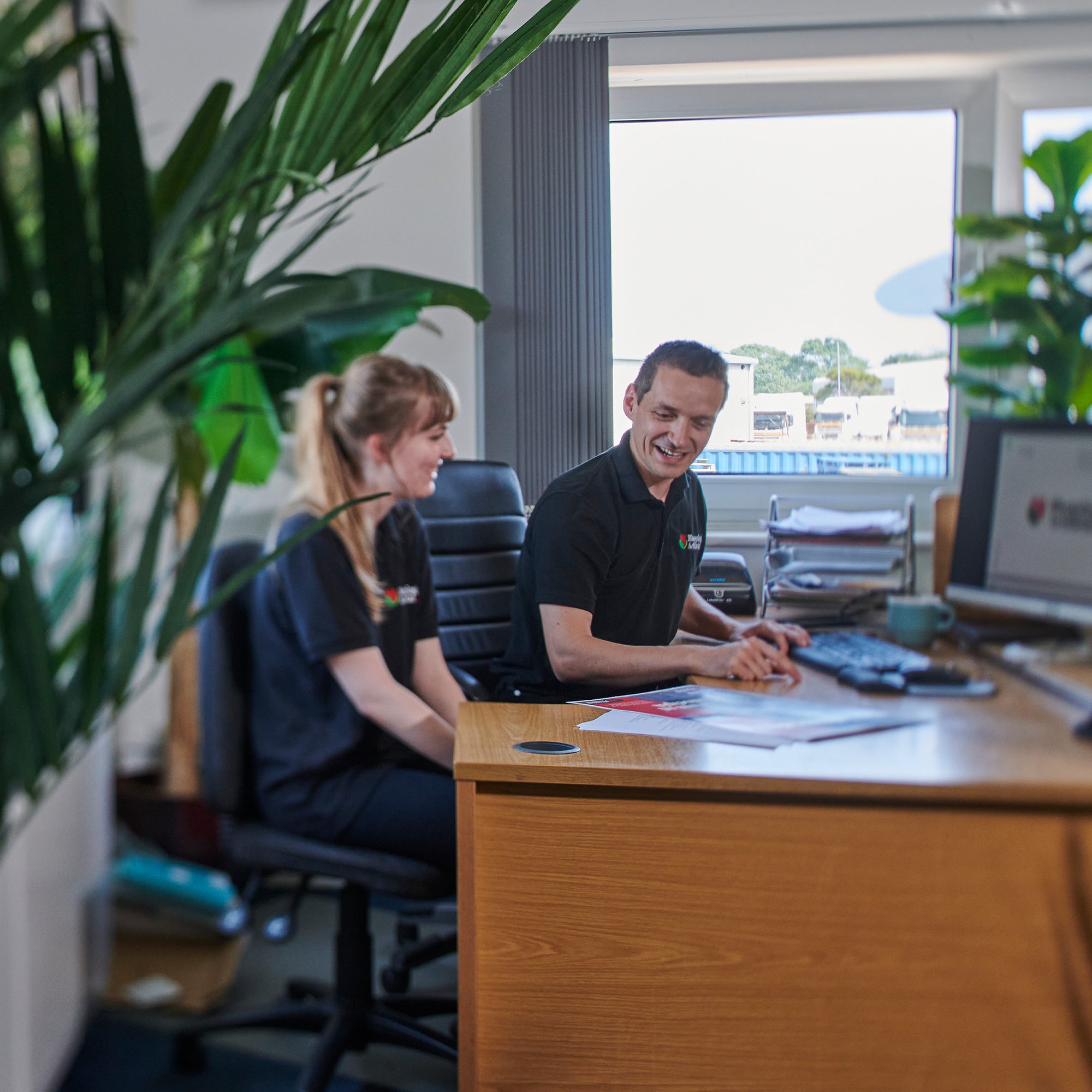 Two people working together at a desk in an office setting with plants and a window in the background.