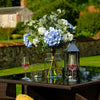 Outdoor setting with a table featuring a vase of flowers, glasses, and a lantern in a garden.