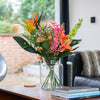 Floral arrangement in a glass vase on a wooden table with a couch and window in the background