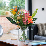 Floral arrangement in a glass vase on a wooden table with a couch and window in the background