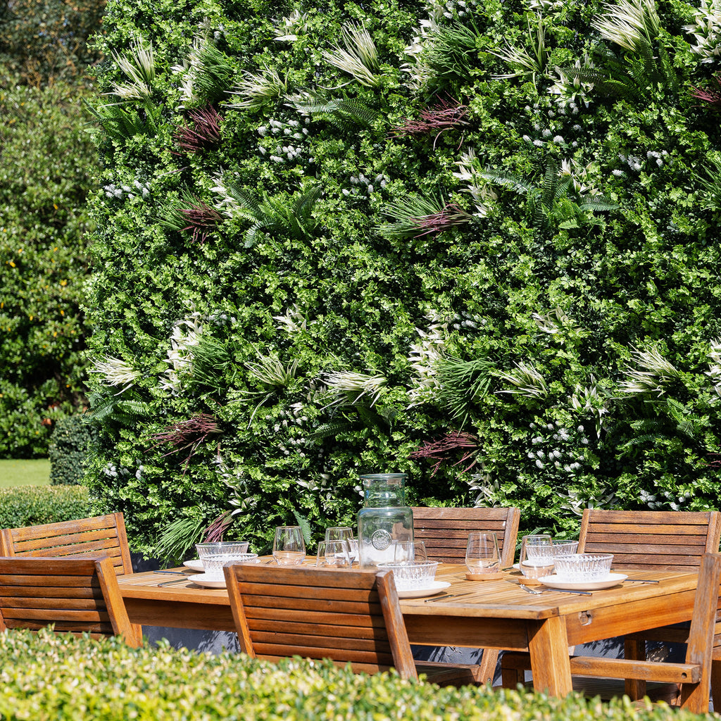 Wooden outdoor dining set with chairs against a lush green wall.