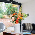 Floral arrangement on a wooden coffee table in a living room with a couch and window in the background.
