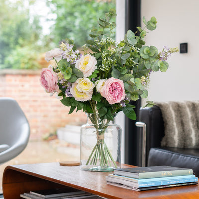Floral arrangement in a vase on a coffee table with books and a couch in the background.