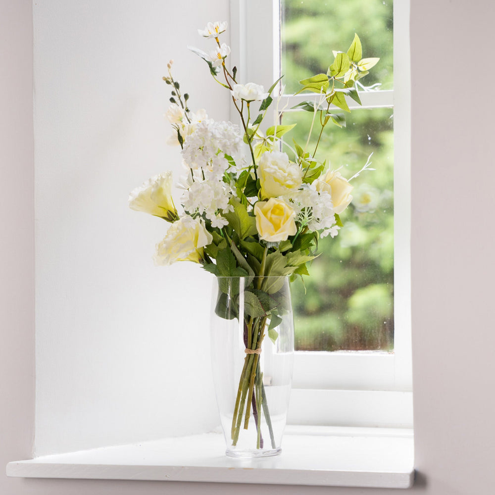 Bouquet of white and yellow flowers in a clear vase on a windowsill.