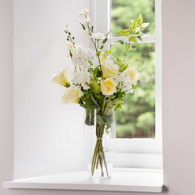 Bouquet of white and yellow flowers in a clear vase on a windowsill.