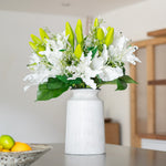 White vase with green and white flowers on a table in a room.