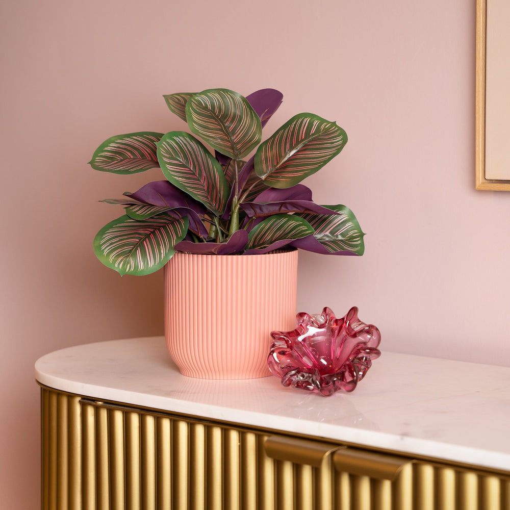 Potted plant and pink glass vase on a marble surface with a gold cabinet and pink wall in the background